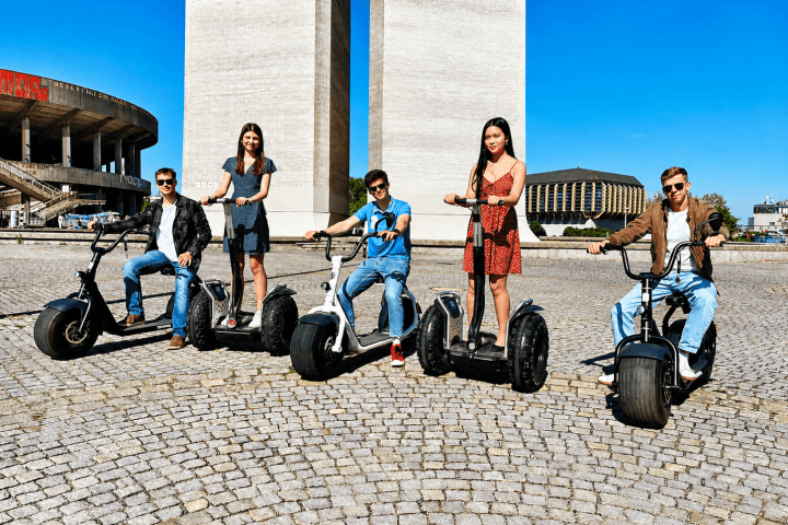 Group of five people on stand-up scooters on cobblestone plaza, tall structures in background, blue sky.