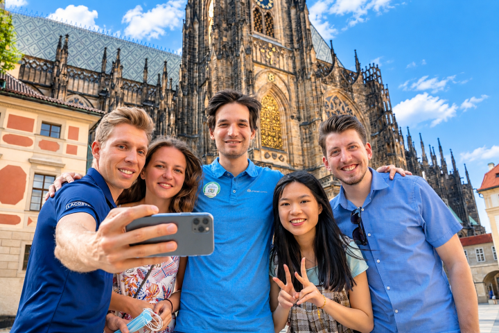 Group of five smiling friends taking a selfie in front of an ornate historic building.