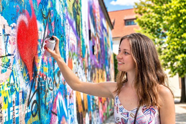 Smiling woman spray-paints a red heart on a colorful graffiti wall.