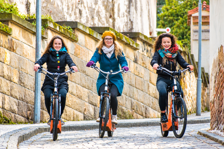 Three women riding bicycles on a cobblestone path, smiling.