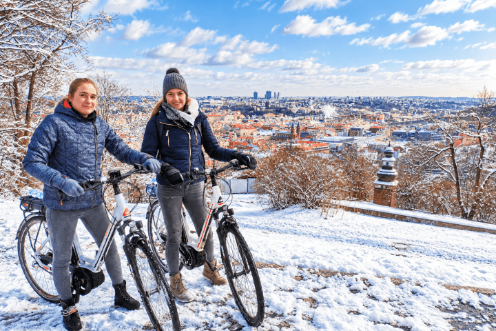 Two people with bicycles in a snowy landscape overlooking a city skyline.