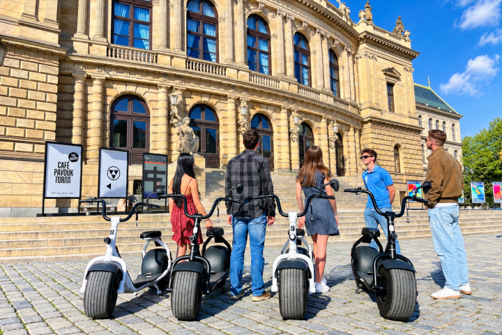 Group with scooters in front of a historic building on a sunny day.