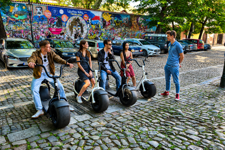 Group on e-scooters talking on cobblestone street near graffiti wall.