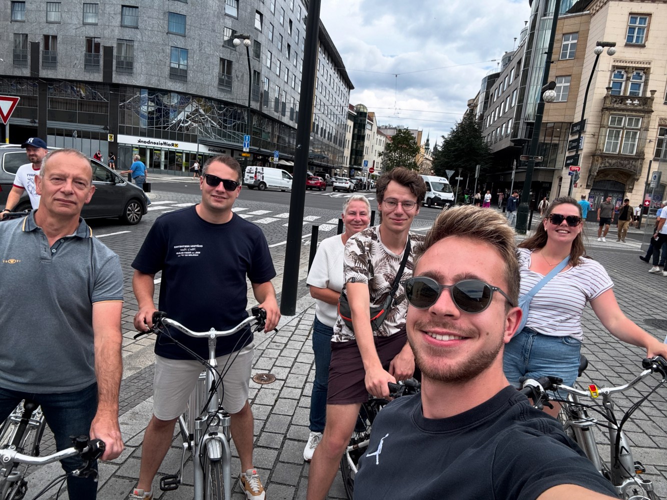 Group of six people with bicycles posing on a city street, surrounded by buildings and traffic.