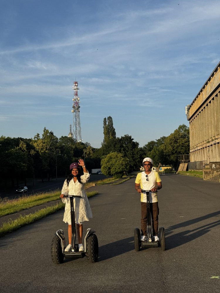 Two people riding Segways on a road, with trees and a tall tower in the background.
