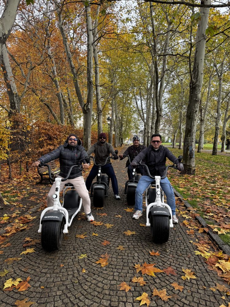 Four people on scooters in a tree-lined path covered with autumn leaves.