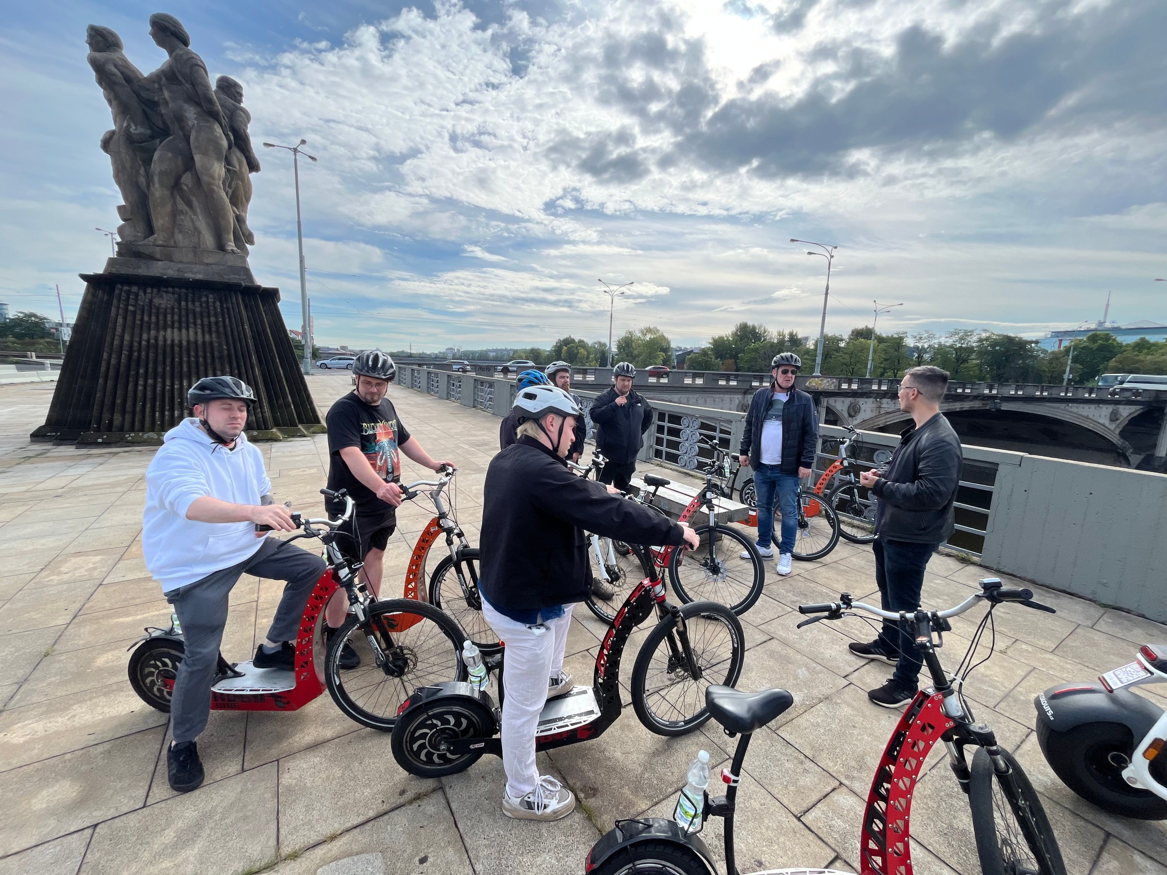Group of people on electric scooters gathered on a bridge with a statue nearby.