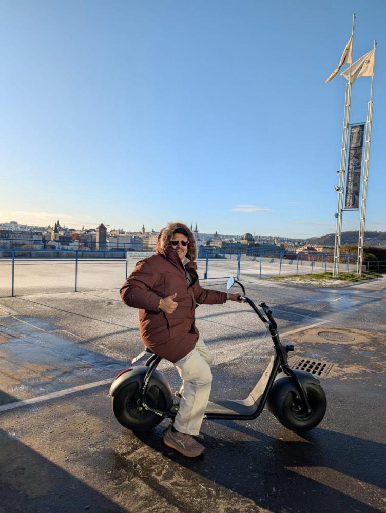 Person in a brown coat rides a scooter, giving a thumbs-up with a city skyline in the background.