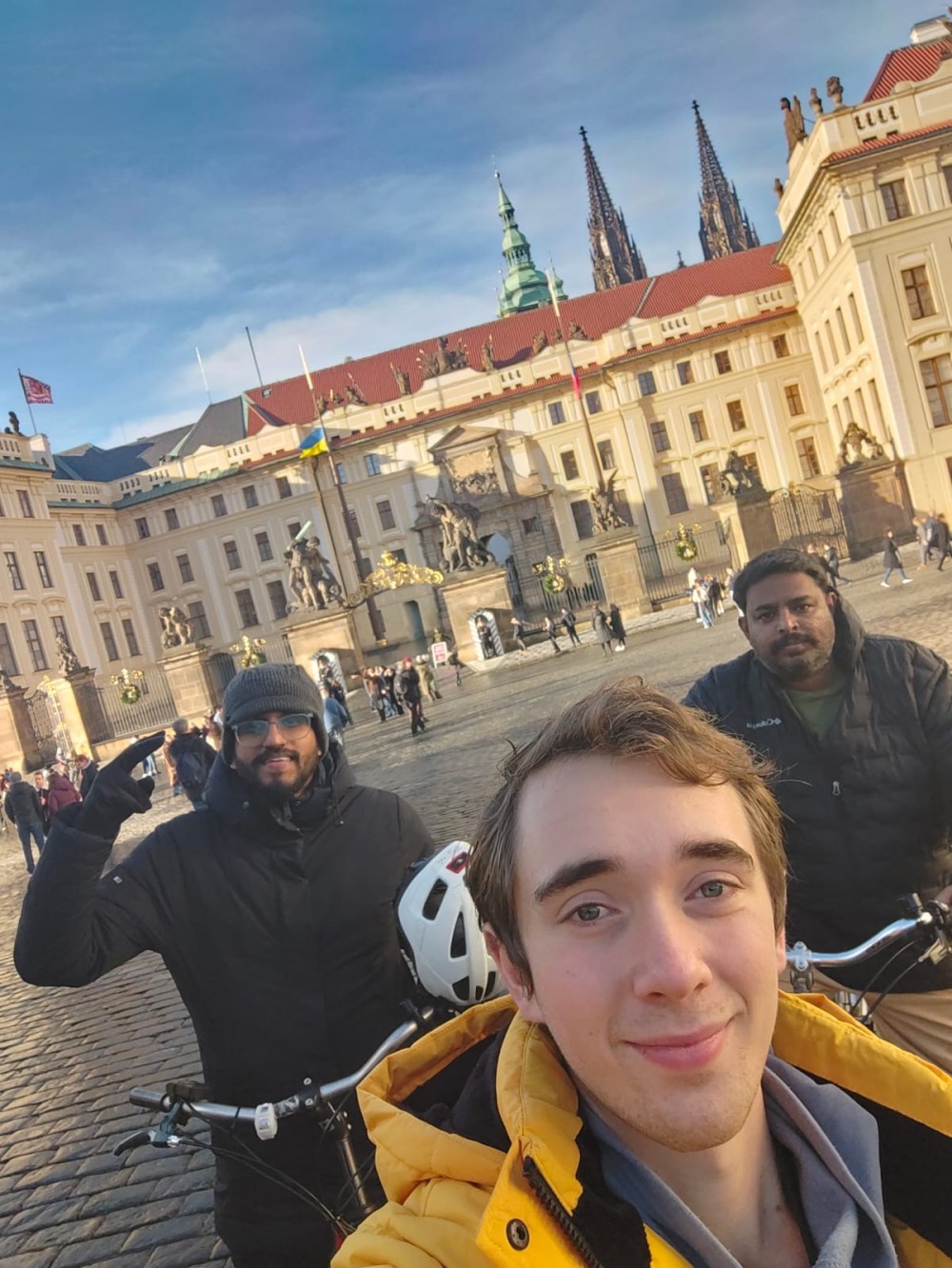 Three people with bikes in front of a historic building with spires on a sunny day.