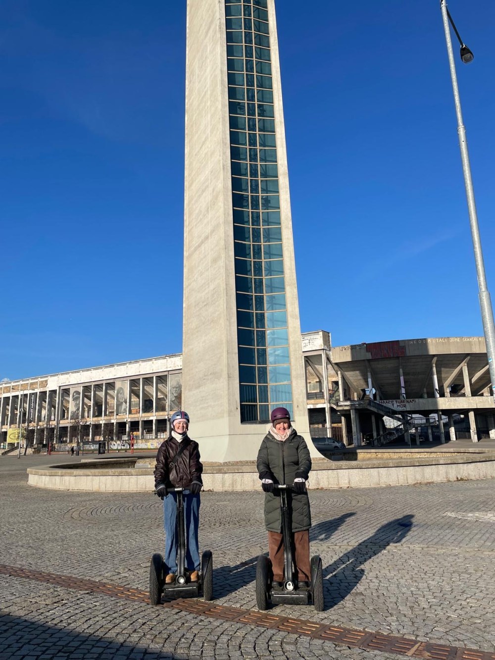 Two people on segways in front of a tall concrete building under a clear blue sky.