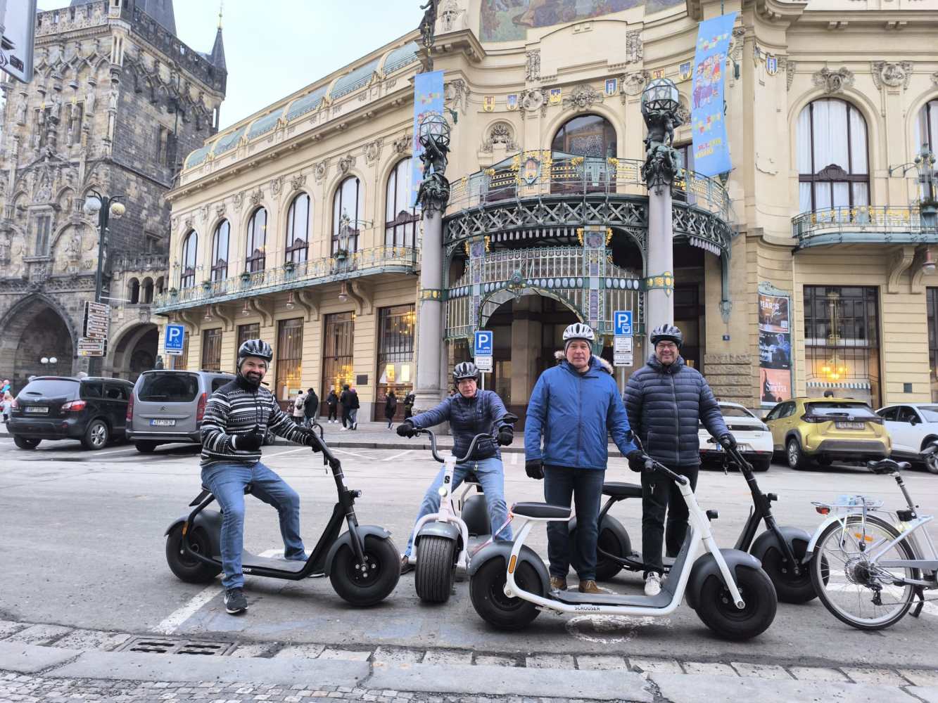 Four people on electric scooters in front of ornate building with arch and tower.