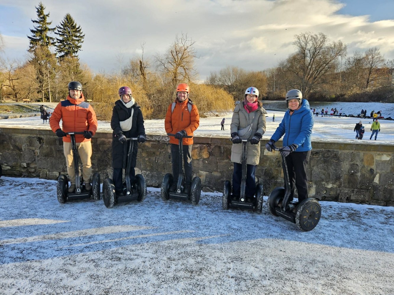 Five people in winter gear on Segways by a snowy park with trees and ice skating in the background.
