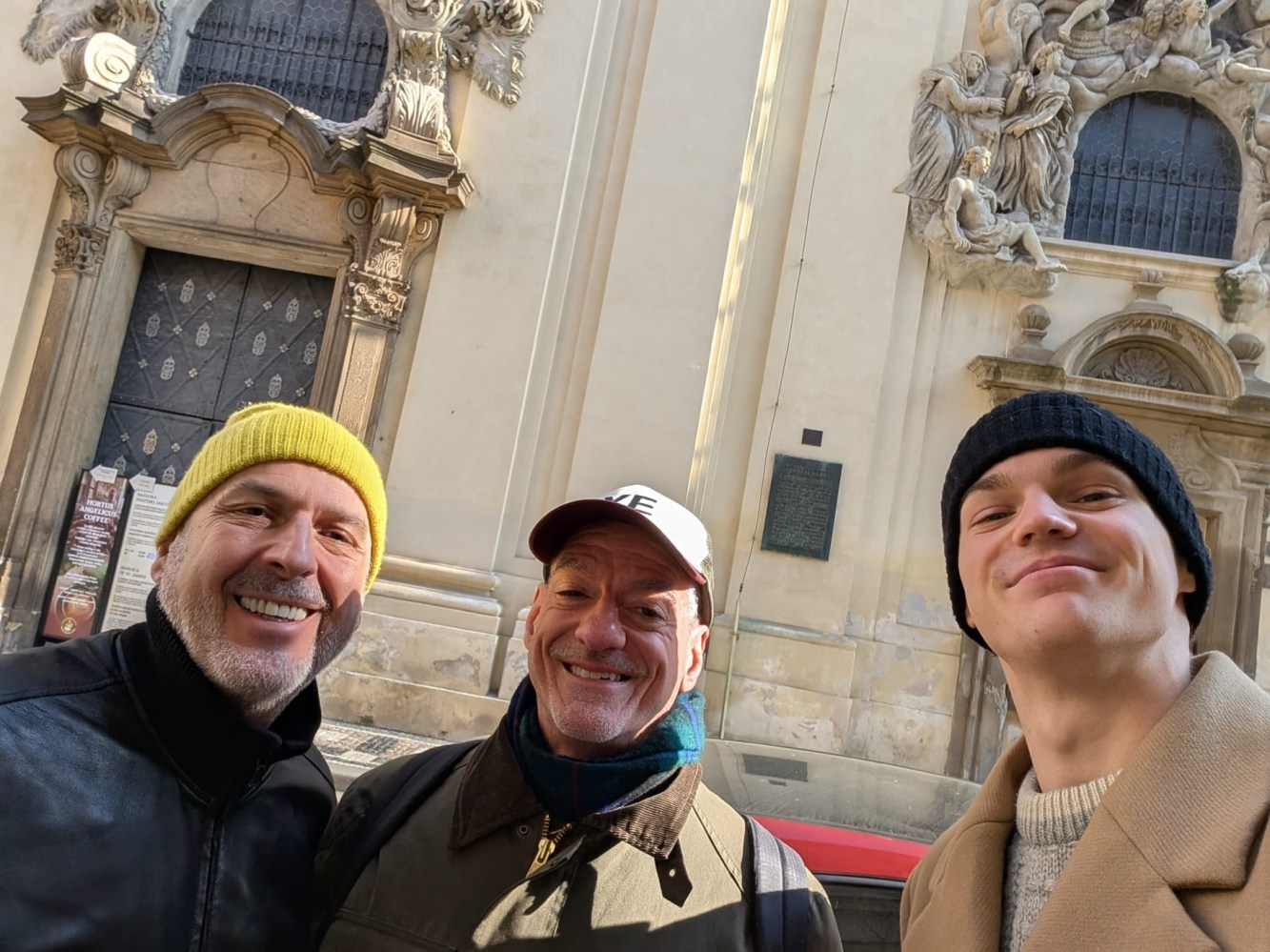 Three smiling men in hats pose for a selfie in front of an ornate building entrance.