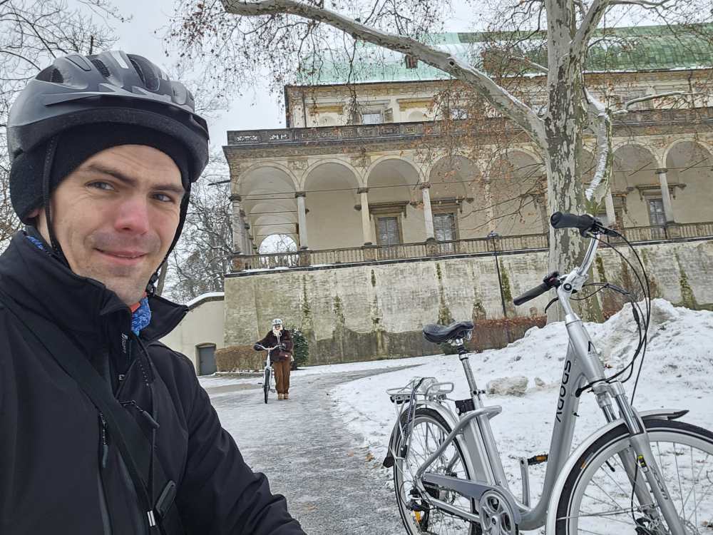 Man in helmet with bicycles in snowy park, historical building in background.