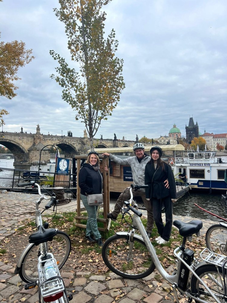 Three people with bikes by a river, bridge, and buildings in the background.