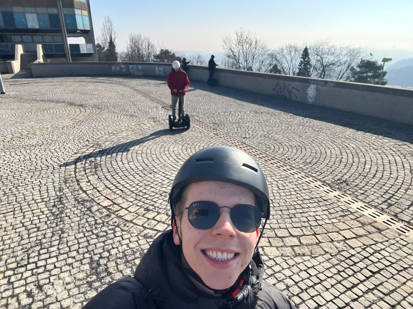 Person in helmet taking a selfie on a stone plaza with others riding electric scooters.