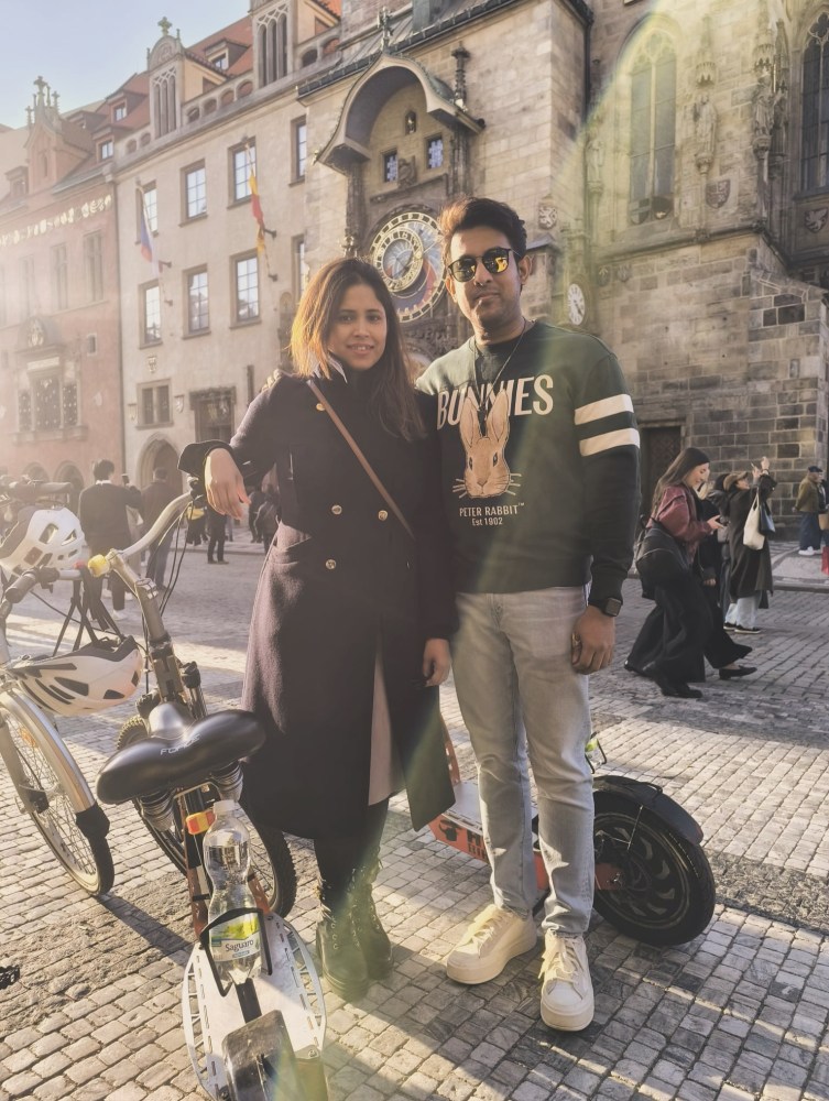 Two people standing with bikes in front of a historic building with an ornate clock on a sunny day.
