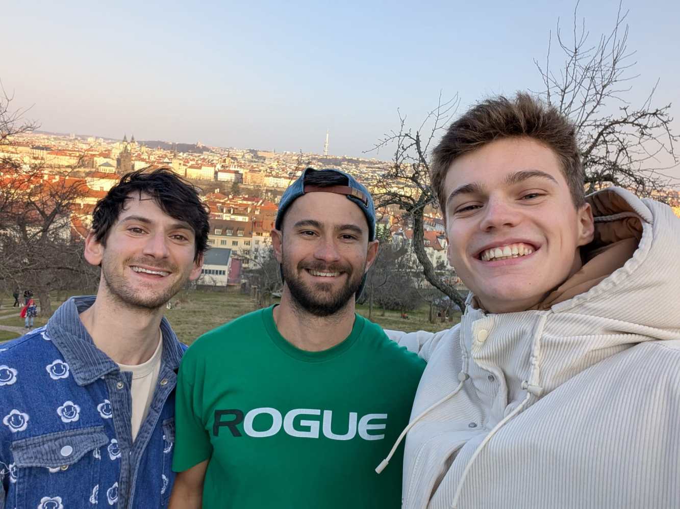 Three men smiling, taking a selfie with a city view in the background.