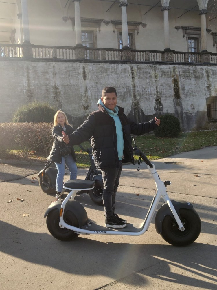Two people on electric scooters smiling in a sunlit park.