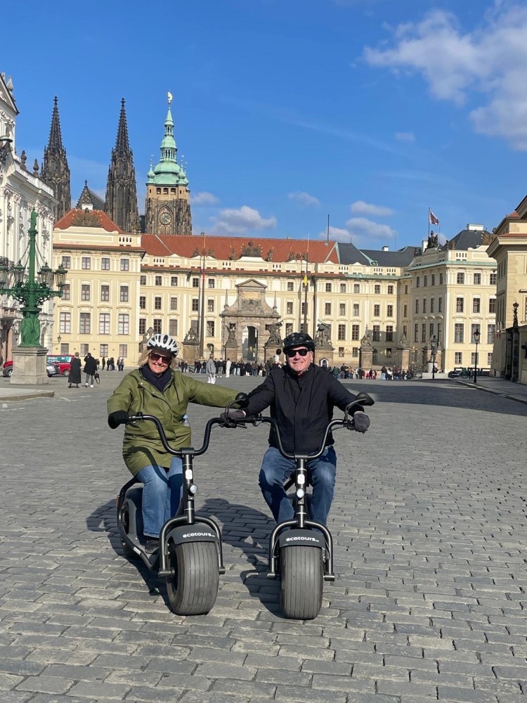 Two people riding scooters in a historical city square.
