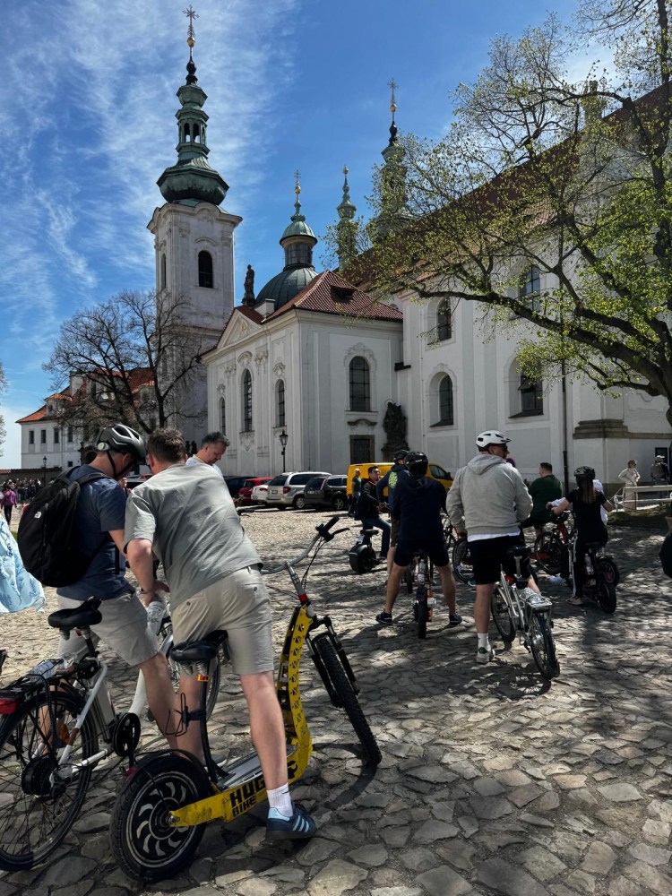 Cyclists gather near a historic white church with tall spires on a sunny day.