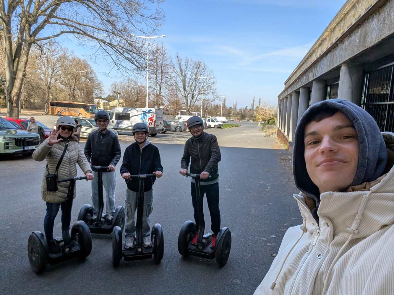Group of four people posing on Segways with helmets; person taking selfie on right in a park setting.