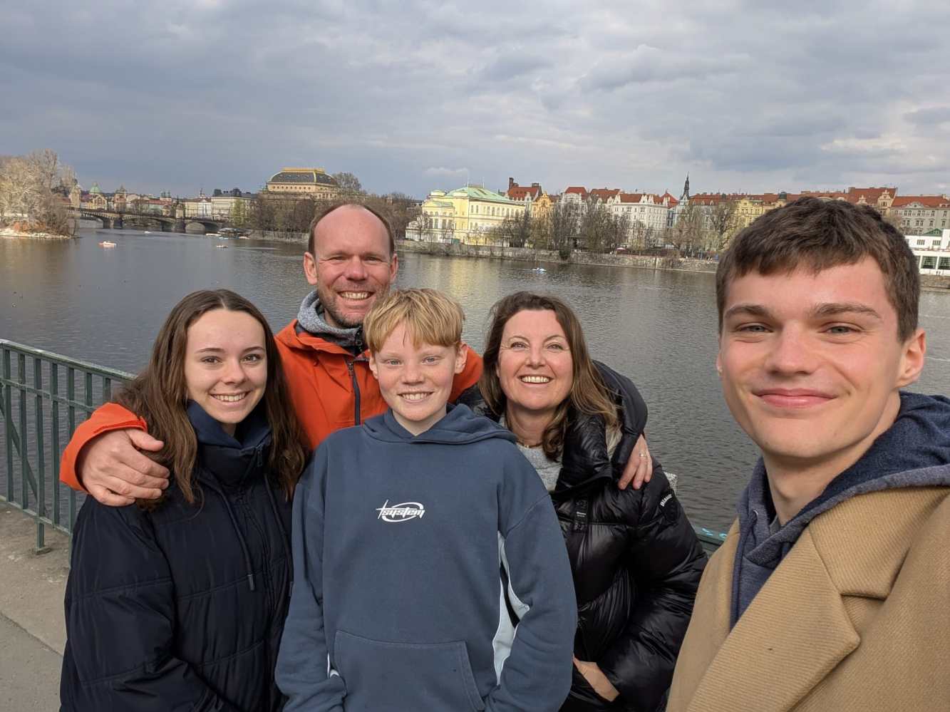 A family of five smiling on a riverside walkway with city buildings in the background.