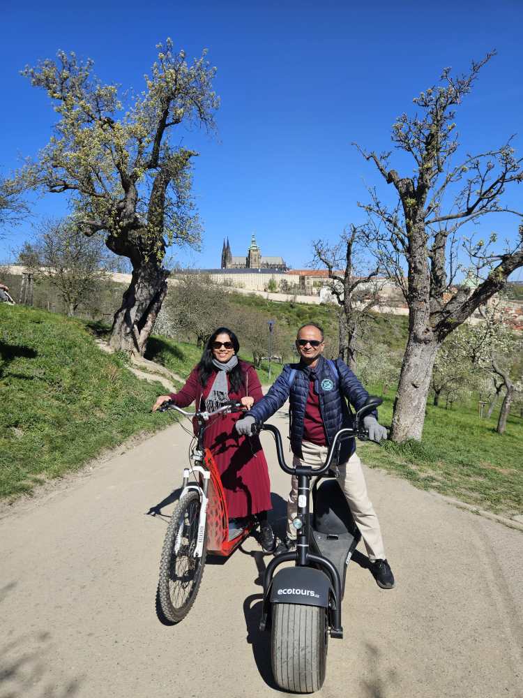 Two people on electric bikes on a path with trees and distant buildings under a clear blue sky.