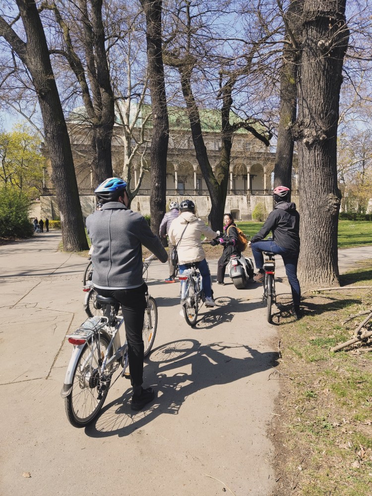 Group of cyclists and a person on a scooter in a park with trees and a building in the background.