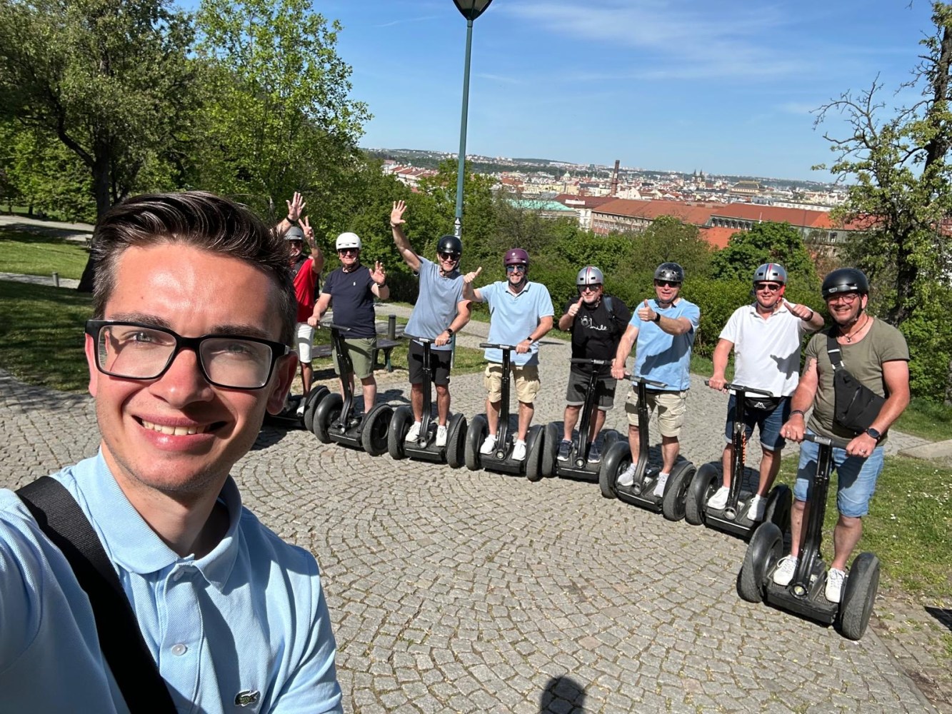 Group of people on Segways pose for a selfie on a sunny day in a park with a city view in the background.