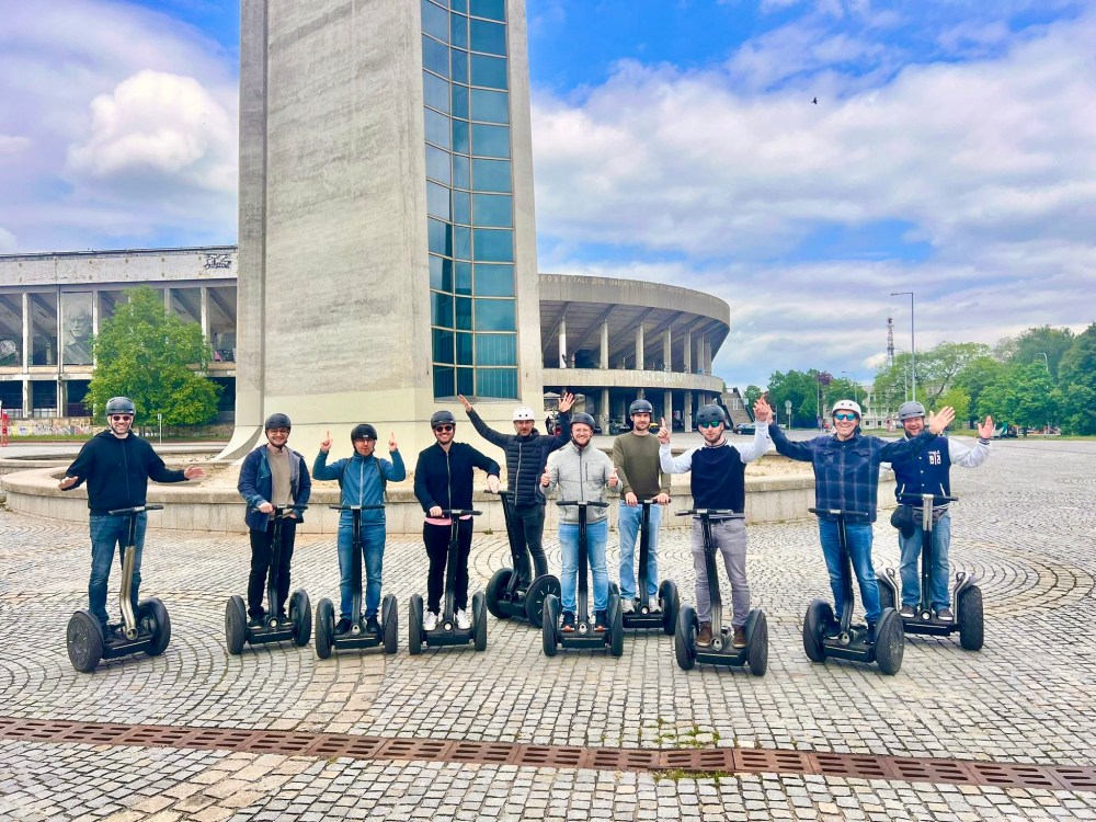 Group of people on segways posing outside a tall concrete structure.