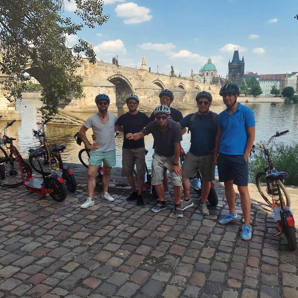 Group of men with helmets standing by electric scooters near a stone bridge.