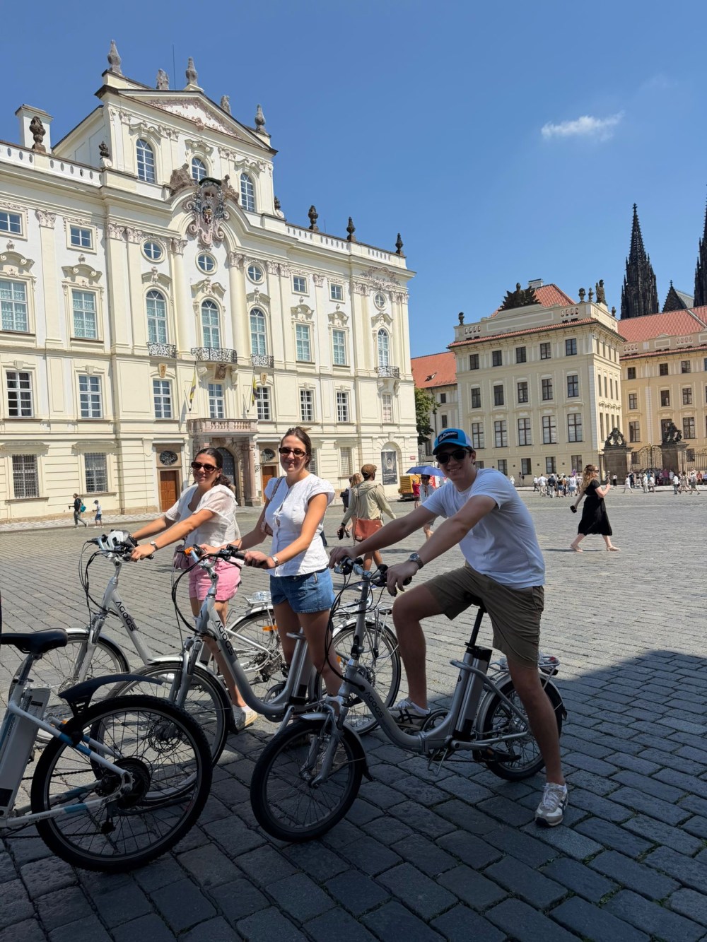 Three people on bikes in front of ornate historic buildings on a sunny day.