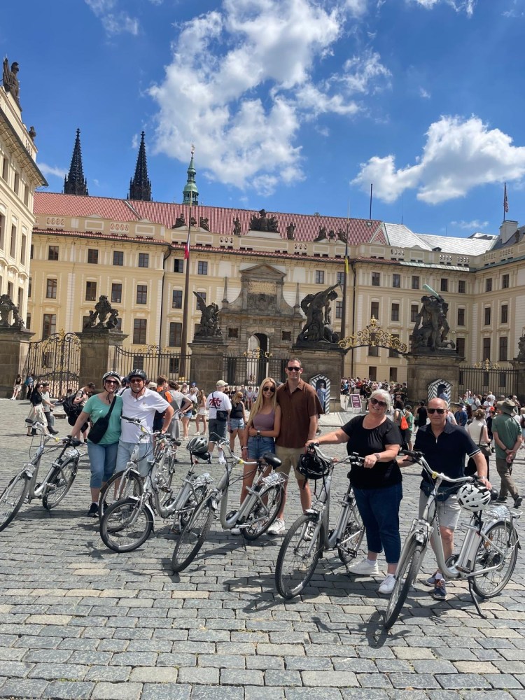 Group of people with bikes in front of a historic building with spires under a blue sky.