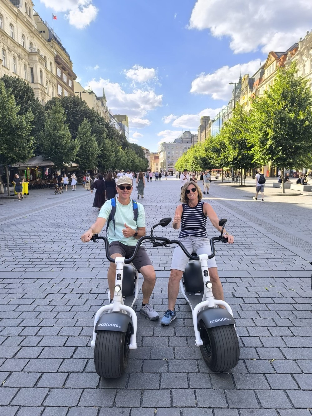 Two people on scooters in a sunny, tree-lined urban street.