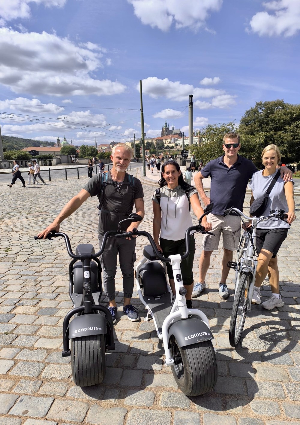 Four people standing with scooters and a bike on a cobblestone street.