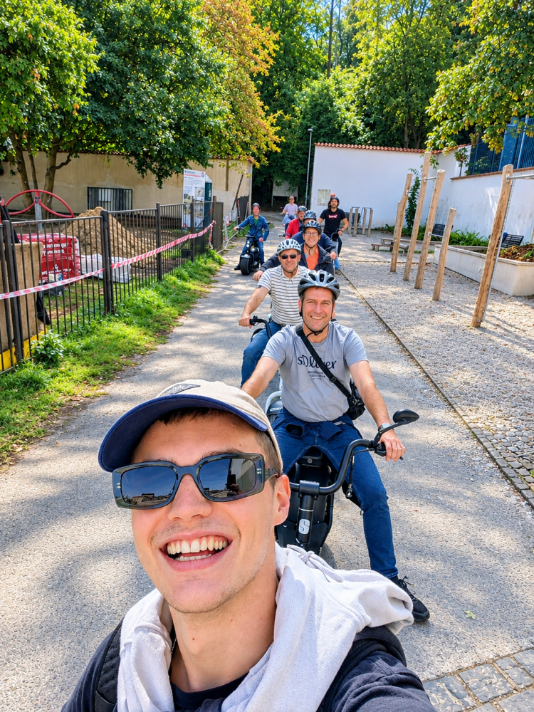 Group of smiling people on scooters, wearing helmets, on a sunny pathway with trees.