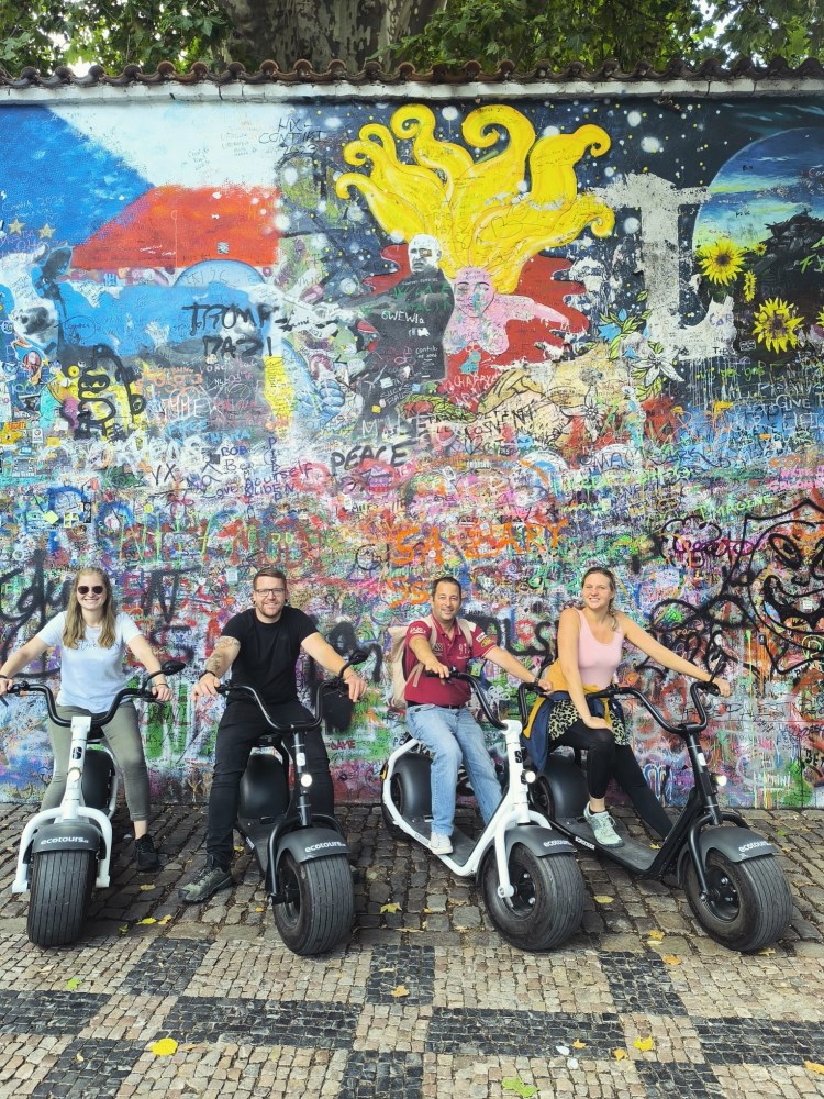 Four people on scooters in front of a colorful graffiti wall.